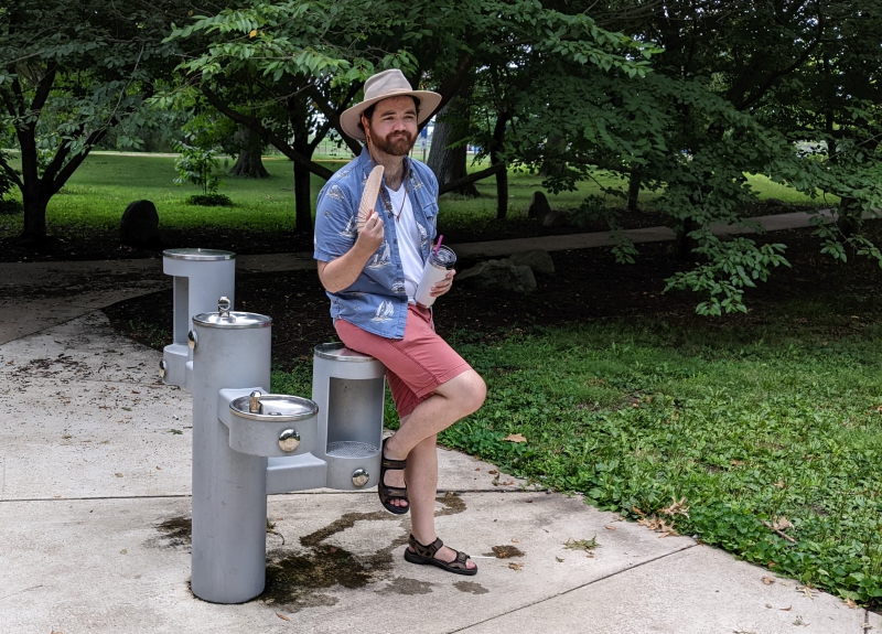 A man in a beige hat, white t-shirt, blue collared shirt, and red shorts is perched on an outdoor water fountain. Photo by Andrea Black. 
