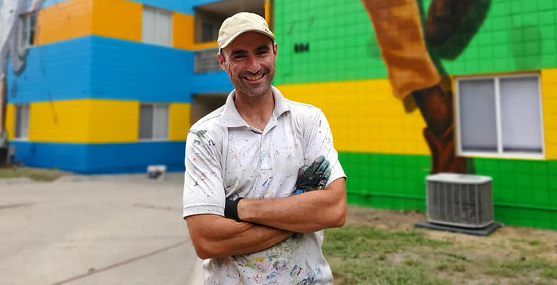 Muralist Rafael Blanco in front of the mural location. Brightly colored stripes cover the building.