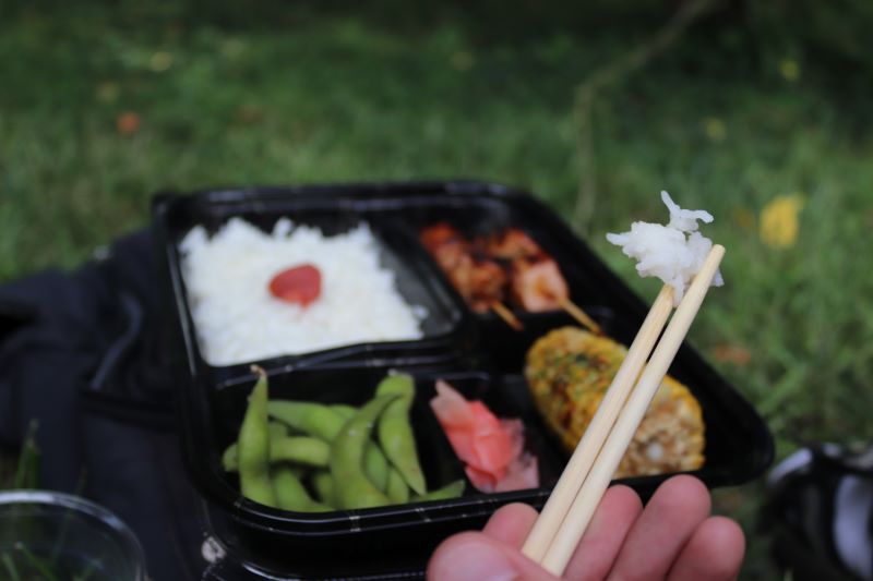 A close up of chopsticks holding a cluster of white rice. In the background is a black bento box with rice, chicken skewers, edamame, and corn on the cob. Photo by Maddie Rice.