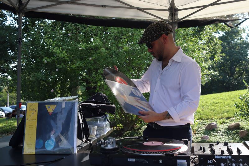 A man in a gray hat, sunglasses, and a white button shirt is pulling a record out of its sleeve. He is standing behind a table with a turntable. Photo by Maddie Rice.