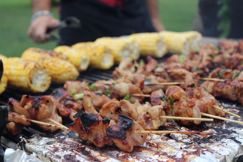 Close up of a large grill with skewers of grilled chicken along the front, and corn on the cob along the back. Photo by Maddie Rice.