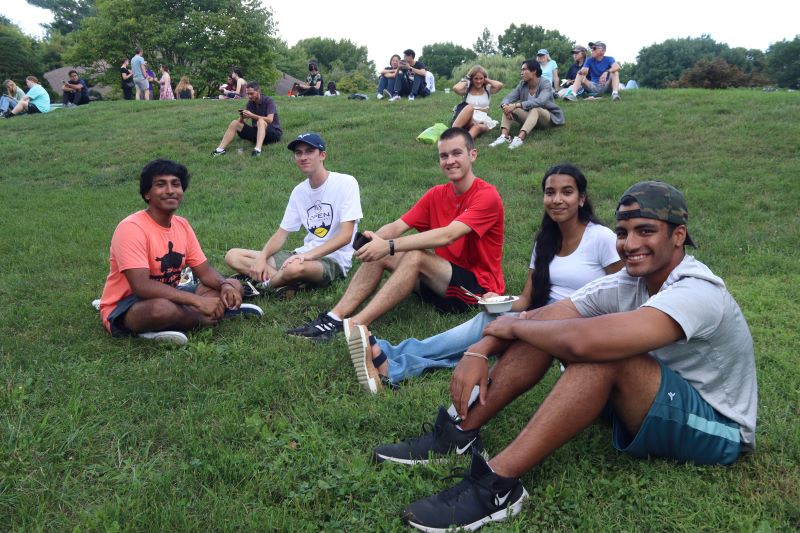 A group of five men and a woman sit together on a grassy hill, looking at the camera. Behind them are several other people gathered in groups. Photo by Maddie Rice. 