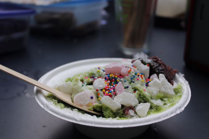A white bowl is filled with shaved ice, colorful sprinkles, and candy pieces. There is a wooden spoon sticking out of the side. Photo by Maddie Rice. 