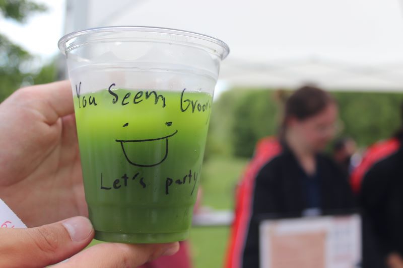 A close up of a clear plastic cup filled with green matcha tea. There is writing in black marker on the cup that says you seem great, let's party. There is also a smiley face on the cup. Photo by Maddie Rice.