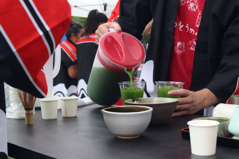 A person in a red t-shirt and black robe is pouring green matcha tea from a clear plastic pitcher into a clear plastic cup filled with ice. There are bowls and a whisk for making matcha sitting on the table. Photo by Maddie Rice.