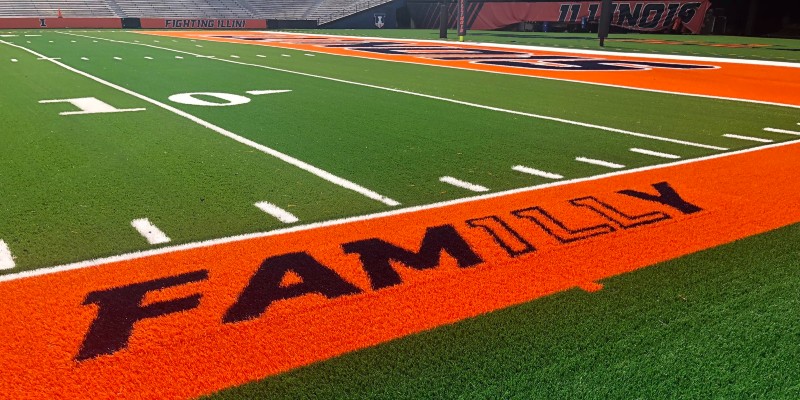 Close up of a turf football field. The sideline is a thick orange line with the word FAMILLY in dark blue. There are white hashlines and yardlines.