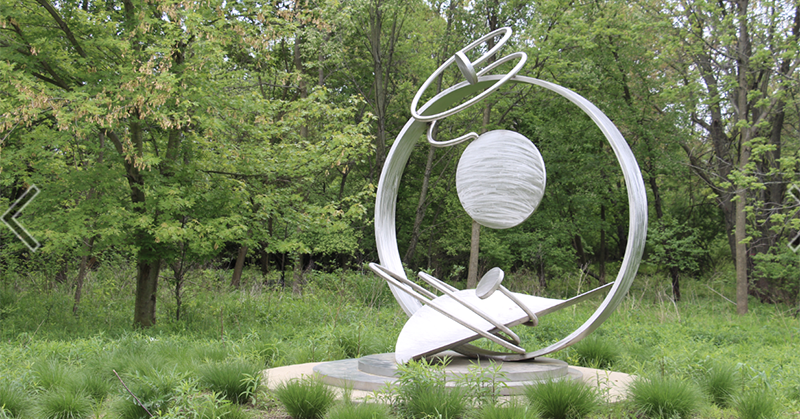 Silver metal circular sculpture with outer rings moving in different directions installed at Meadowbrook Park. 