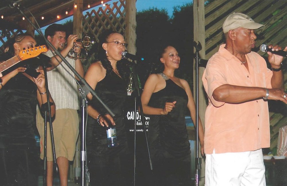 Musician Candy Foster sings on stage, with background singers behind him, as well as a trumpet player.