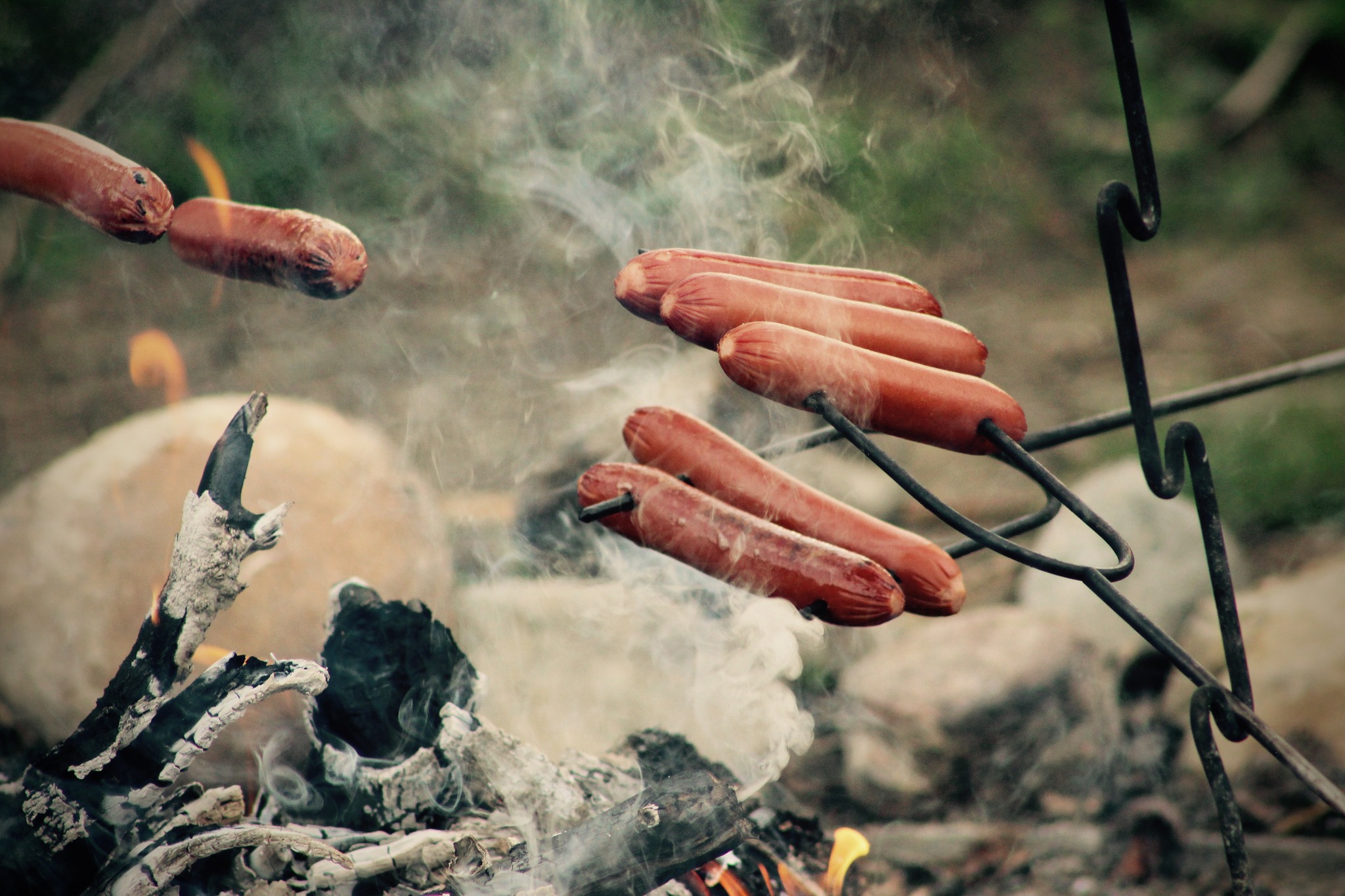 Hot dogs are speared on roasting tools over a firepit. The background is blurred out, but looks like rocks and grass. Photo from Sleepy Creek Vineyard Wieners & Wine Facebook event page.