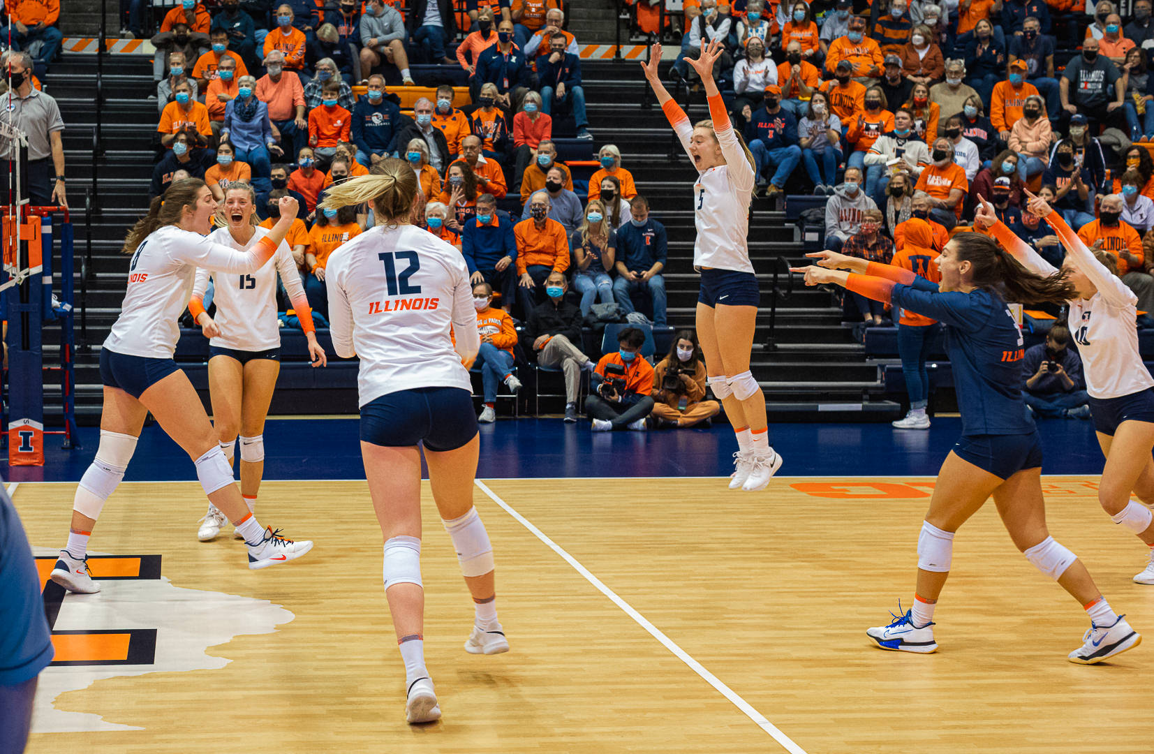 The Illini Womenâ€™s Volleyball team celebrates after winning a point or a game. On the left, three tall, white women in white jerseys move toward the right, where a tall, white woman with a long blonde ponytail is jumping in the air celebrating. On the right, there are two more women pointing in celebration. Both are white; one wears a white jersey, one wears a blue jersey. Photo from the Facebook event page. 