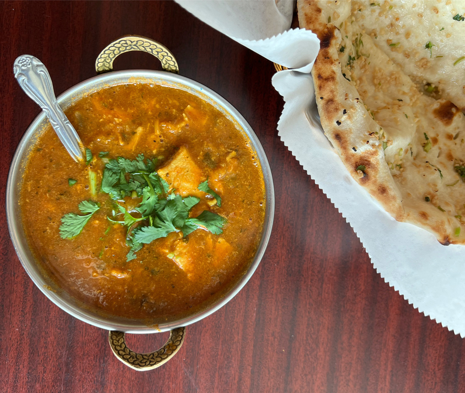 On a brown table, there is a brass bowl of chicken curry beside a basket of garlic naan. Photo by Alyssa Buckley.