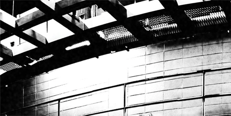 Black and white noir-style photo of open ceiling grid with a view of brick wall in the background.