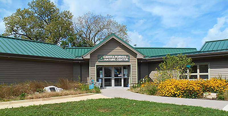 A wide shot of the front of the Anita Purves Nature Center in Urbana. A low tan building with green roof and details, sitting on a grass lawn with trees in the background.
