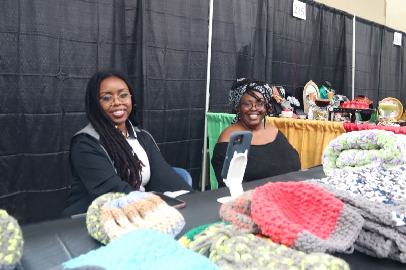 A Black woman with glasses and long braids, and a Black woman with glasses and a colorful headdress sit behind a table filled with knit hats and blankets. Photo by Maddie Rice.