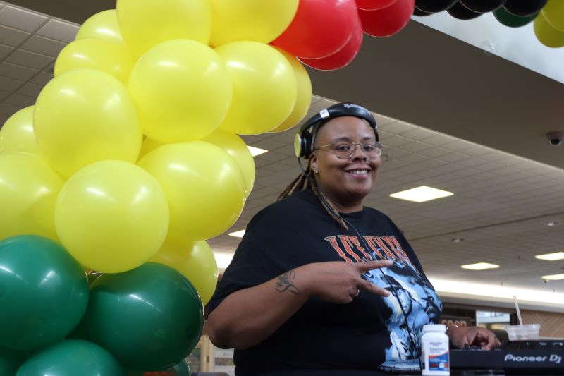 A Black woman with headphones, glasses, and a black t shirt stands in front of a balloon arch made with black, yellow, and red balloons. Photo by Maddie Rice.