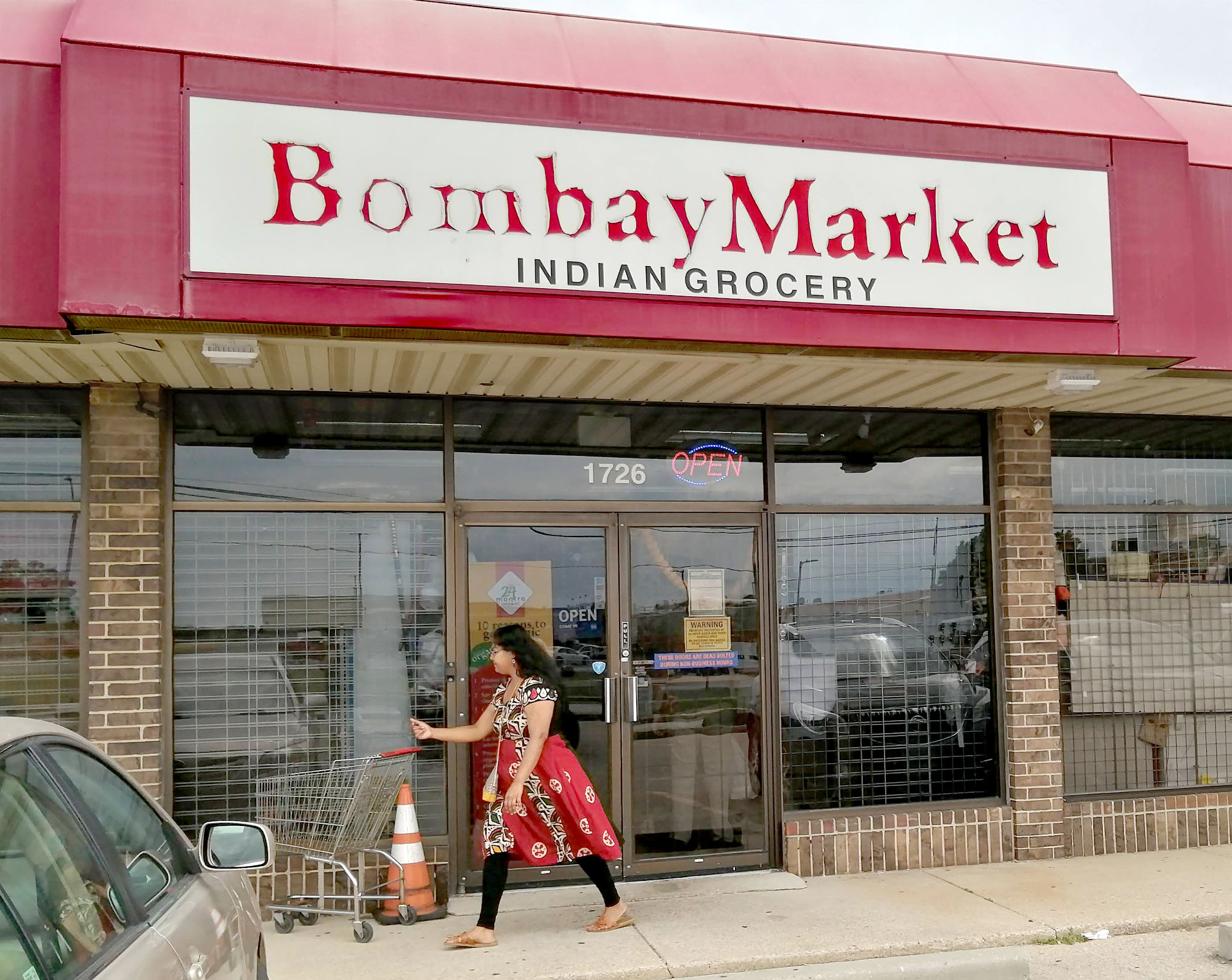 A storefront in a stripmall with a shopper walking by; a sign prominently displays â€œBombay Market Indian Grocery.â€ Photo by Paul Young.