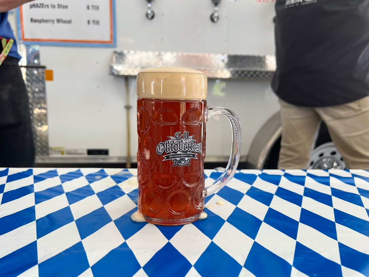 A clear beer stein filled with a dark amber colored lager and a head of foam sits on a table with a blue and white patterened tablecloth.
