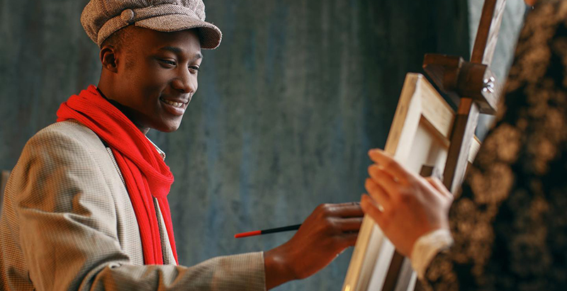 Photo of Black painter working at a standing easel holding a brush dressed in a tan coat, red scarf and brown tweed cap, with a Black woman facing him with her hand on the back of 