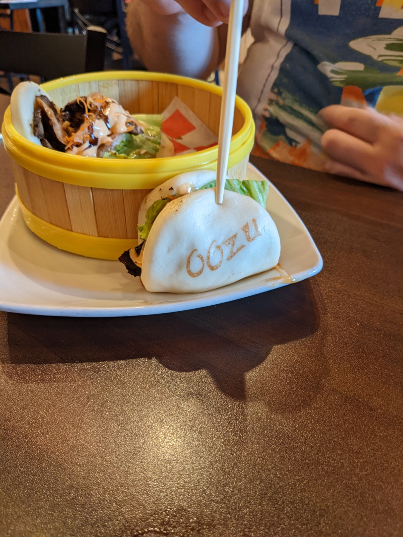 A square white plate holds a wooden steamer basket. On the plate in front of the basket is a steamed bun with an â€˜Oozuâ€™ branding. In the basket is a second bun, slightly opened, Lettuce, carrots, pork, and sauce can be seen inside. Photo by Caitlin Aylmer.