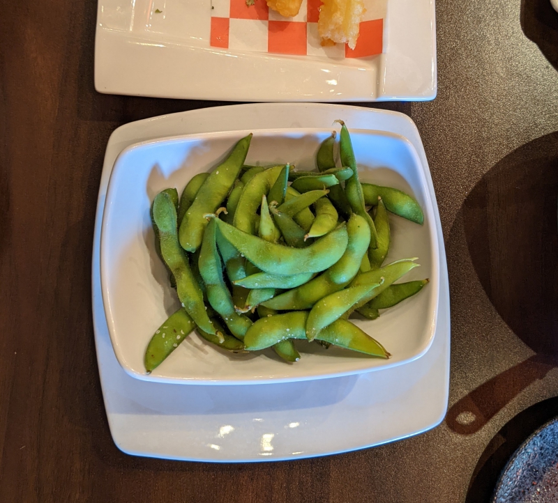 A white rectangular bowl sits on a white square plate. The bowl is filled with green edamame pods. Photo by Caitlin Aylmer.