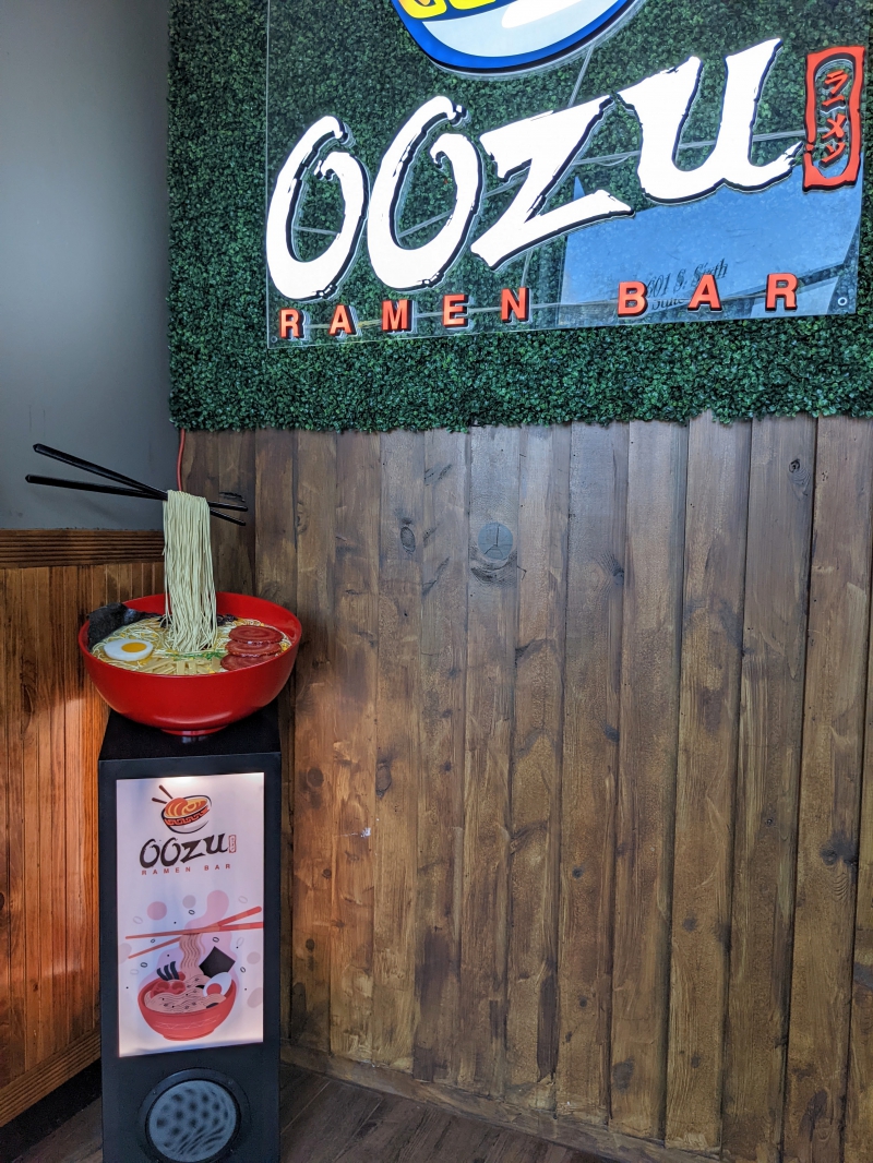 Inside the vestibule, a statue of a large red bowl is prominently on display in the corner. A pair of chopsticks hold ramen noodles above the bowl. On the far wall is a trendy display with a sign reading â€˜Oozu Ramen Bar.â€ Photo by Caitlin Aylmer.