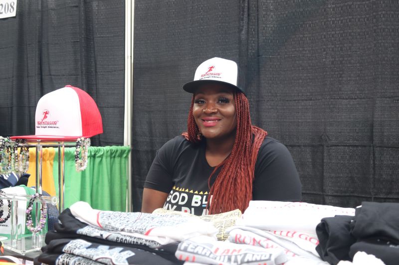 A Black woman with long braids, wearing a black and white trucker hat and black t shirt, sits behind a table filled with folded shirts. Photo by Maddie Rice.