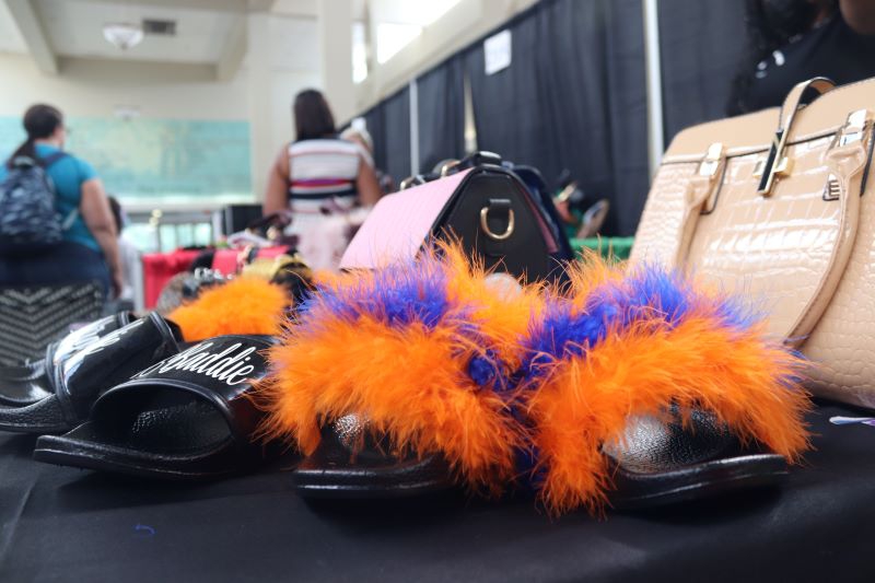 A row of decorated slide sandals, some black with writing and others with orange and blue poofs, sit on a table in front of a row of purses. Photo by Maddie Rice.