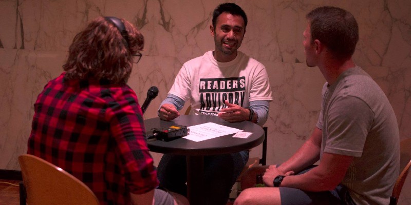A brown-skinned man wearing a white t-shirt with a black and white logo that says "readers advisory." He is talking with two white people who are facing him, their backs to the camera. On the left of the image, the person is wearing a red and black plaid flannel shirt. On the right a white man is wearing shorts and a gray t-shirt. They are sitting around a small round dark metal table.