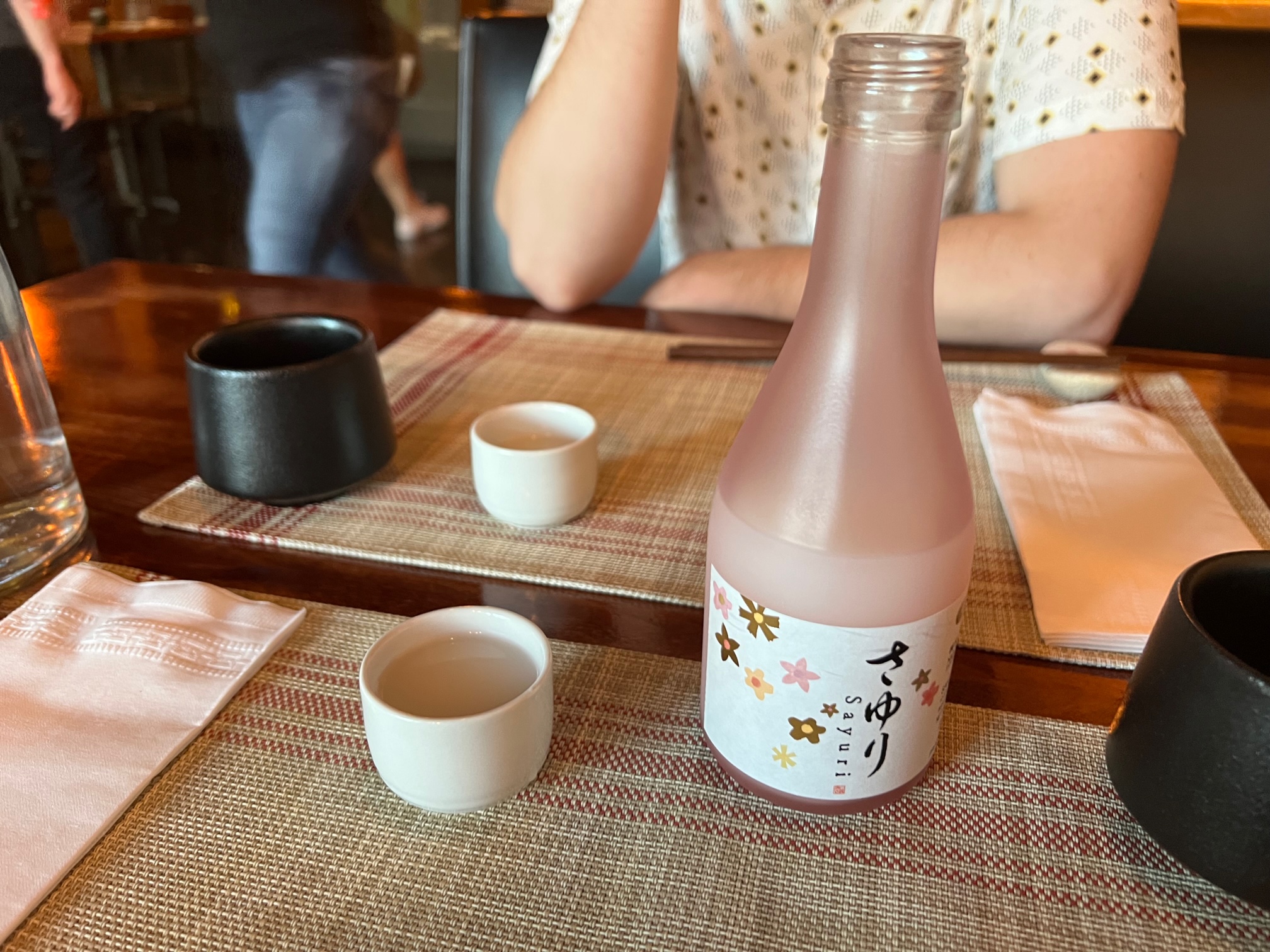 A bottle of chilled Sayuri pink sake sits on a tablemat beside a white sake cup. The author's husband is across the table and just his shirt and arm are visible behind his white cup of sake. Photo by Alyssa Buckley.