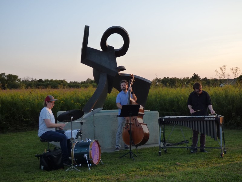 A jazz trio: a drummer, upright bass player, and xylophone player are playing music in front of an abstract statue. Prairie grasses are behind them, and the sun is setting. Photo from Urbana Park District Facebook page.