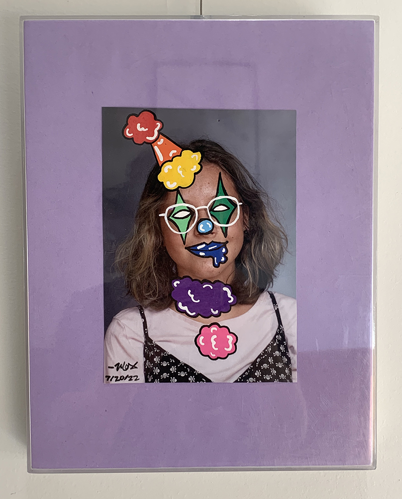 A class photo with a clown face painted over it is mounted in lavender and hung against a white wall.