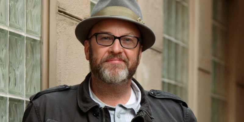 Mark Roberts, a middle aged white man, is pictured from the chest up. He is wearing a dark colored jacket, dark framed glasses, and a gray-green hat. He has a graying beard.