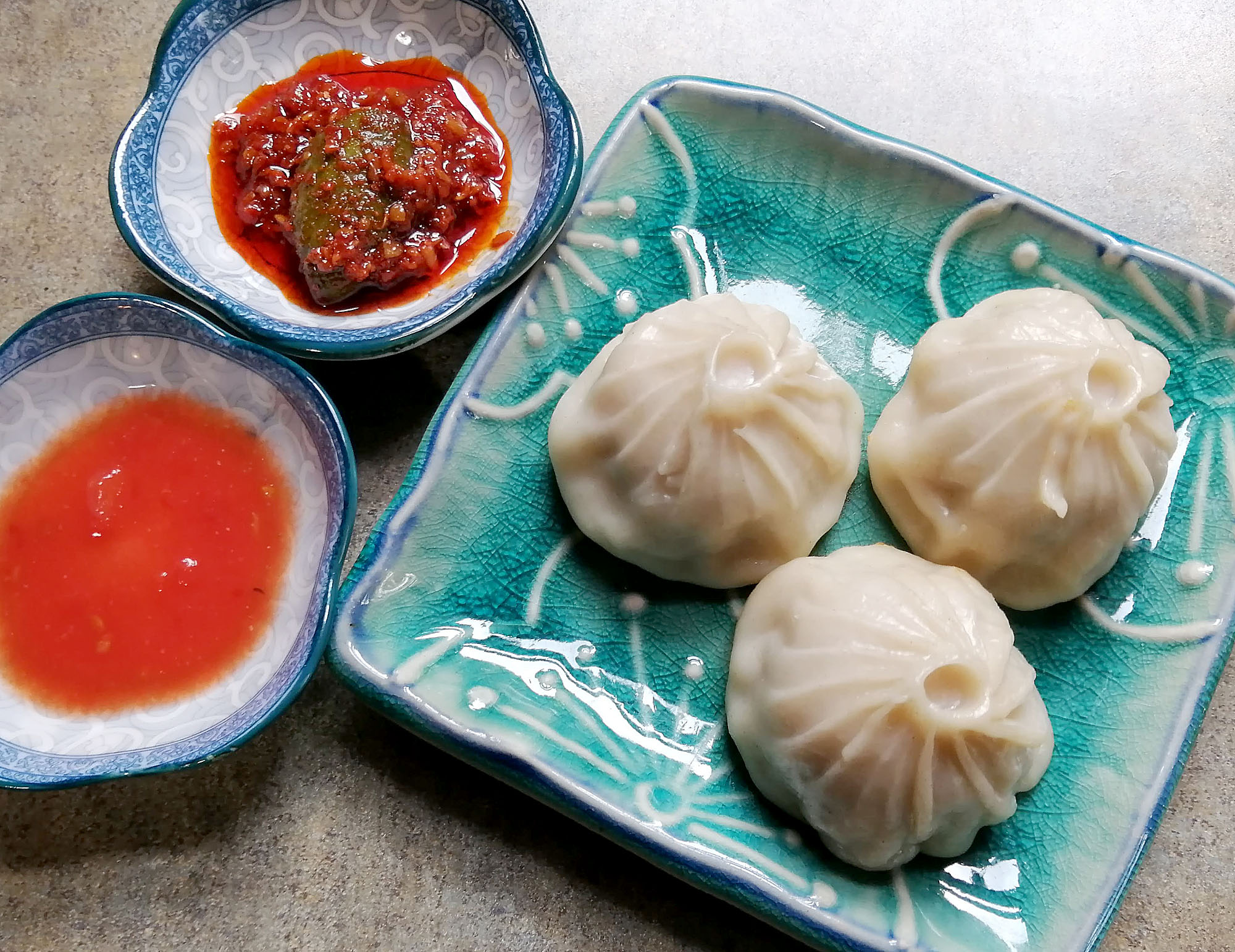 A blue plate with three steamed dumplings, plus two smaller dishes with dipping sauces on the side. Photo by Paul Young.