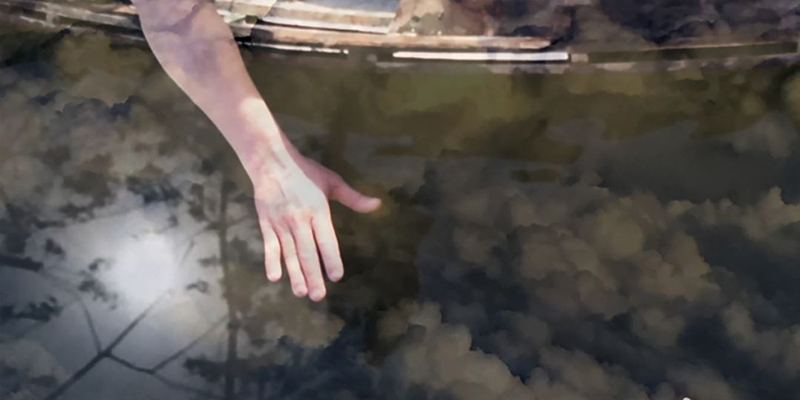 Close up of color photo of hand reaching down into what appears to be water but is in fact sky. 