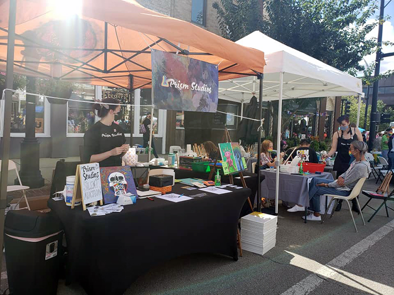 Prism Studios booth from Pride 2021 featuring completed art, art supplies, and groups of people painting.