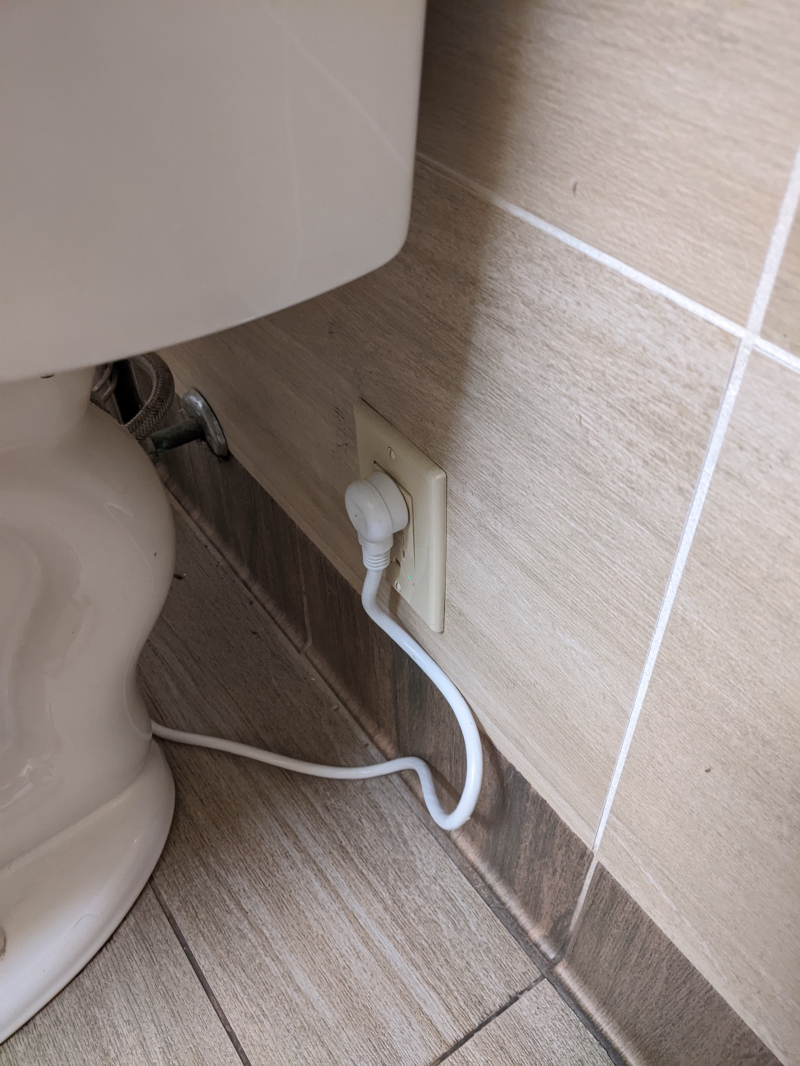 A close up of an electrical outlet behind a white toilet. There is a white cord attached to the toilet that is plugged into the outlet. Photo by Tom Ackerman.