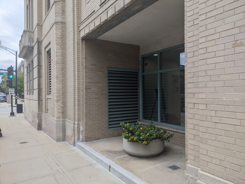 A vestibule recessed from the sidewalk. There are tall windows, and a round concrete planter sits in front of the windows. It has yellow flowers planted in it. Photo by Tom Ackerman.