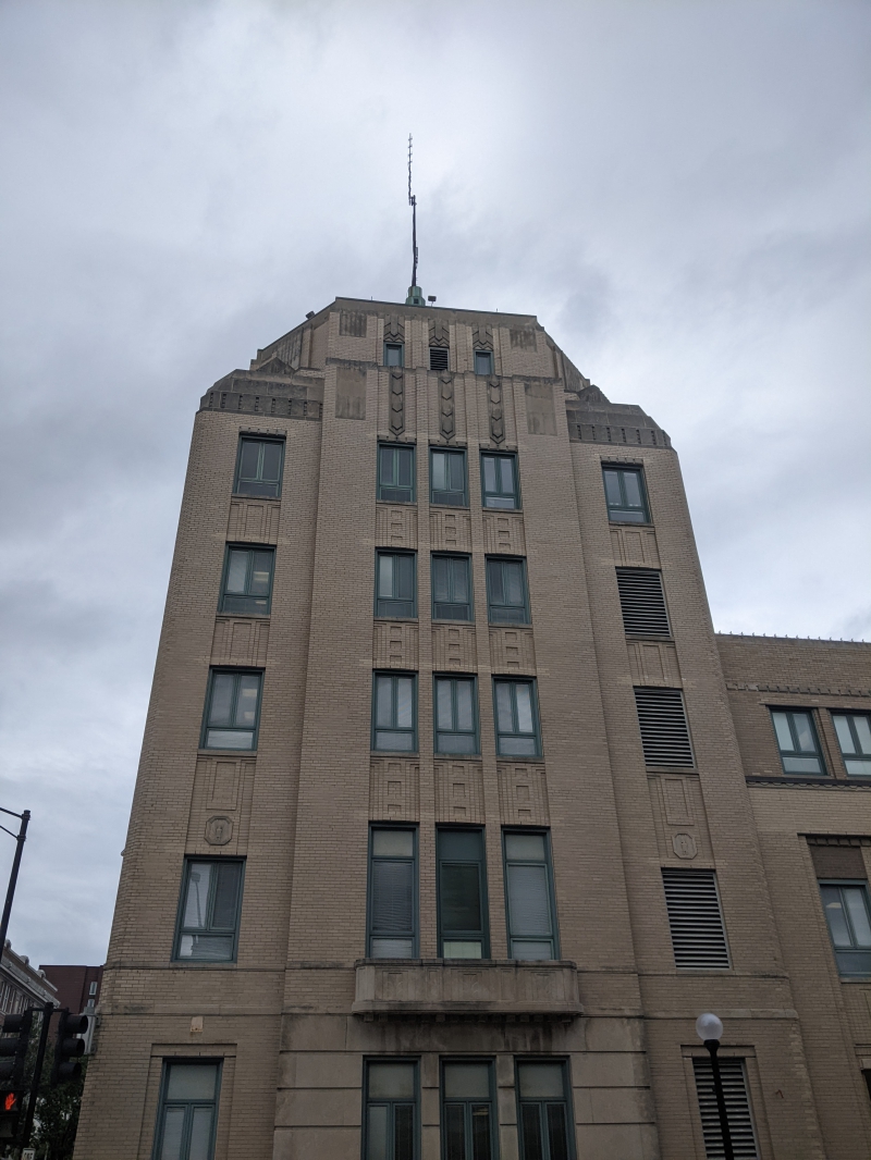 A close up of the 6 story portion of the city building. It has thin rectangular windows, beige brick, and an antenna on the roof. Photo by Tom Ackerman.