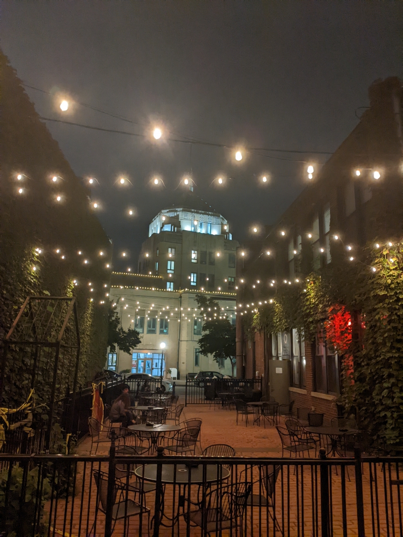 A few of the illuminated city building through beer gardens with black wrought iron tables and chairs. String lights criss cross over the top. Photo by Tom Ackerman.
