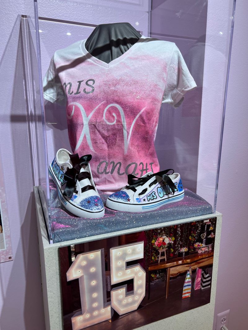 A display featuring an airbrushed t-shirt and sneakers. At the bottom of the display is a photo of a large lighted 15 and a table with flowers and gifts. Photo by Julie McClure.