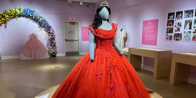 A mannequin is dressed in a red quinceanera dress, dark wig, and tiara. It stands on a wood pedestal at the front of a museum gallery. 