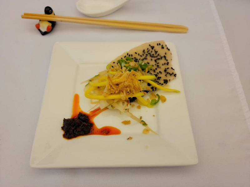 Rice paper salad with the black sesame rice paper sticking out of the salad and the chili oil gathering in the opposite corner of the square plate. Photo by Matthew Macomber.