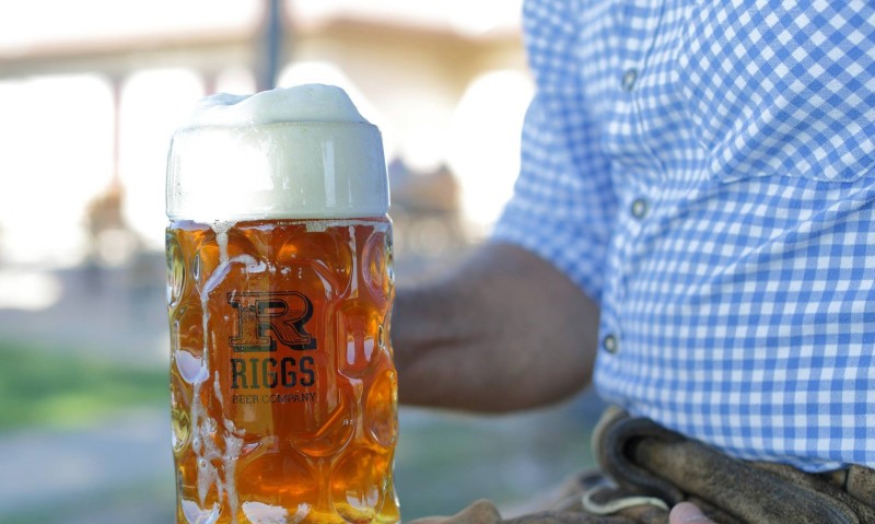 Close up of a glass beer stein filled with amber colored beer, with white foam on top. It sits on the knee of a man in a blue and white checked shirt. Photo from Facebook event page.