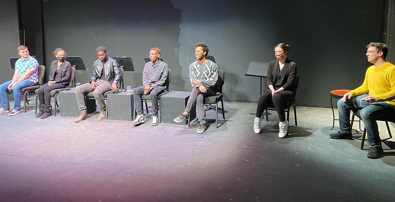 Black and white actors sitting in a semi circle on the Station Theatre stage. 