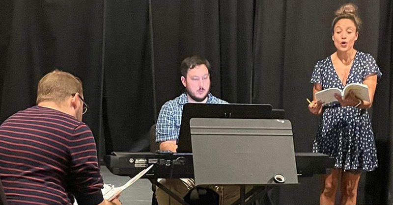 Three cast members at rehearsal at Station Theatre, on left white male is seated with his back to the camera, center is a white male keyboard player and too the right is a white female singer with sheet music. 