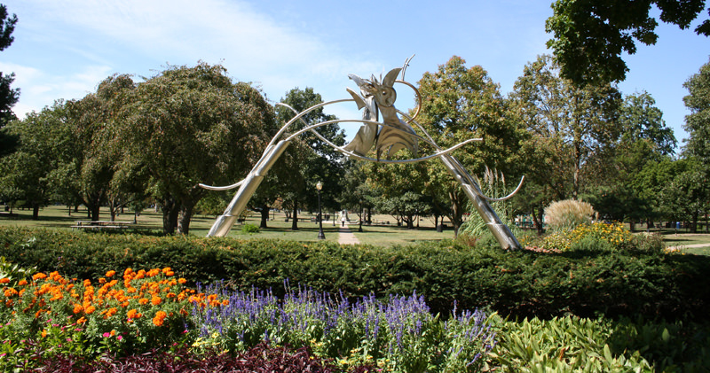 A view of West Side Park in Champaign featuring an arch-shaped silver sculpture, flower beds and trees.