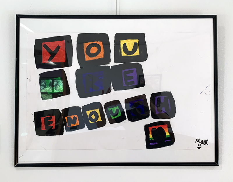 A painting with Pride rainbow color filled black outlined boxes containing the letters Y O U A  R E E N O U G H framed and hung against a white wall.