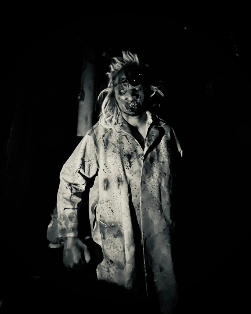 Black and white photo of a person in a bloody lab coat and scary mask with wild hair. Photo from Baldwin Asylum Facebook page.