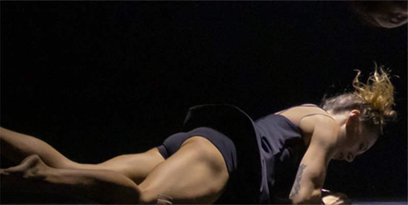 Female performer stretched face down on the stage with legs and feet pointing up. Performer is dressed in form-fitting grey shorts and tank top.