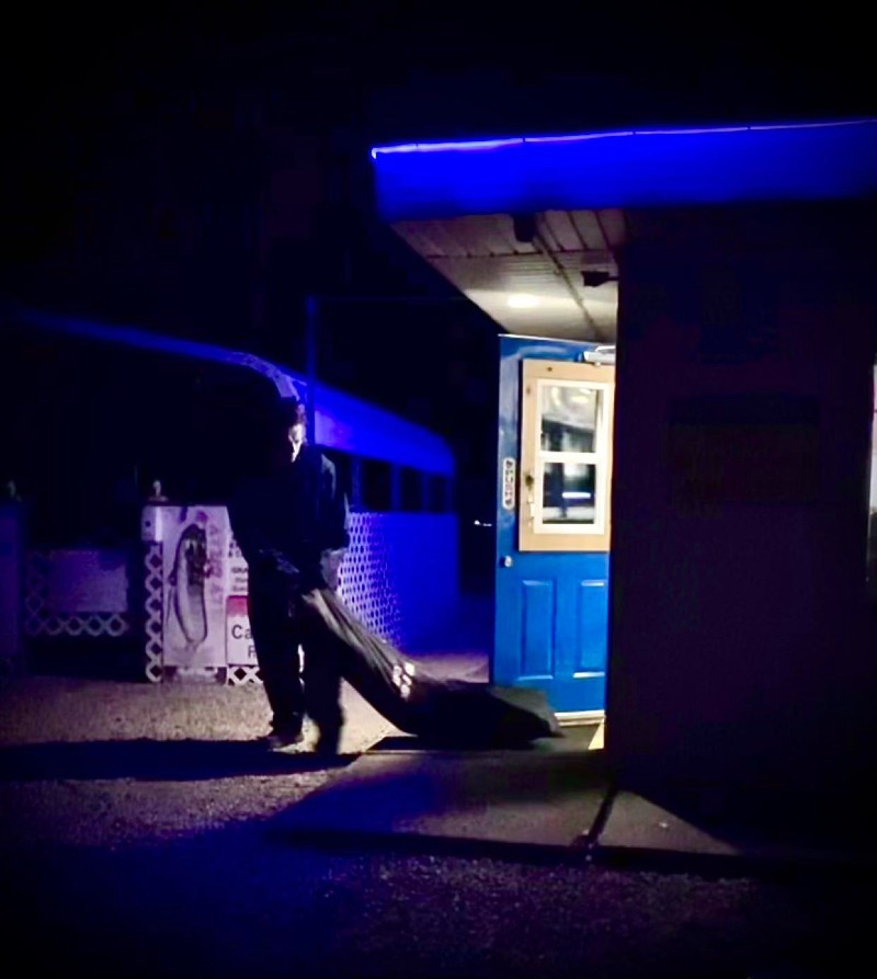 A person dressed like Michael Myers in a white mask and blue jumpsuit is dragging a black body bag from a lit doorway at night. Photo from Harvest Moon Drive In Facebook page.